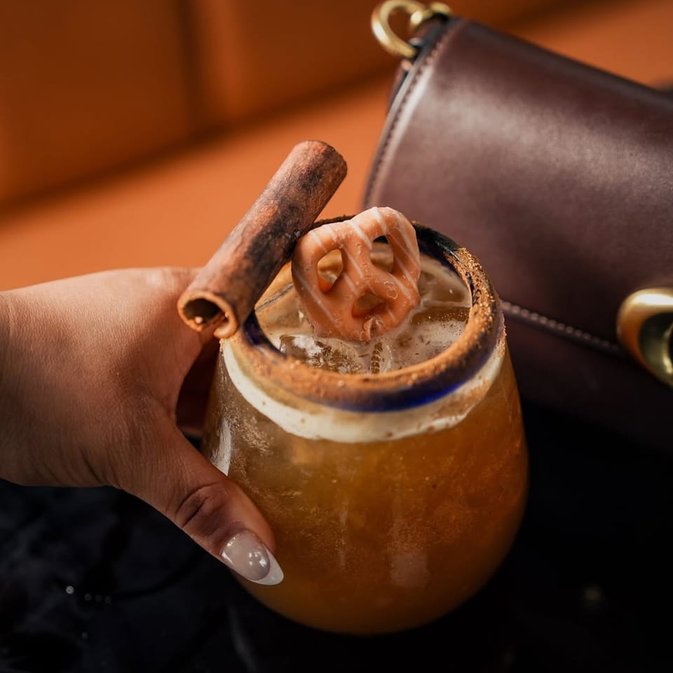 Hand holding a Mezcal cocktail garnished with a cinnamon stick and a small pretzel.