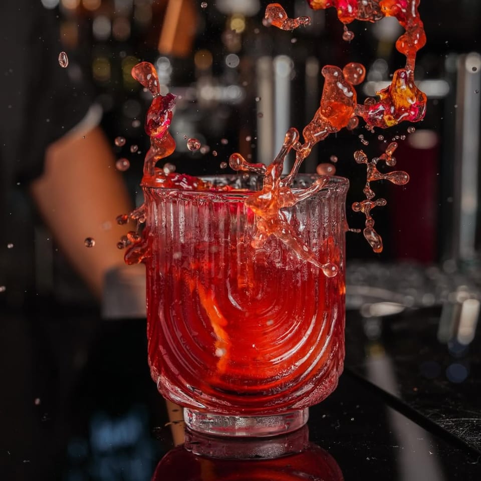 Red cocktail splashing out of a textured glass on a dark bar with blurred background lights.