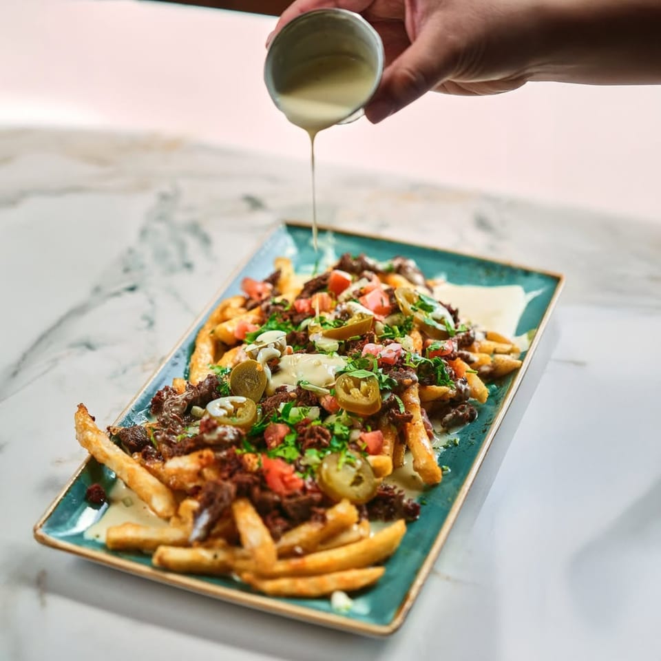 Hand pouring creamy white sauce over Mezcal's Mexican fries topped with meat, jalapeños, and tomatoes.