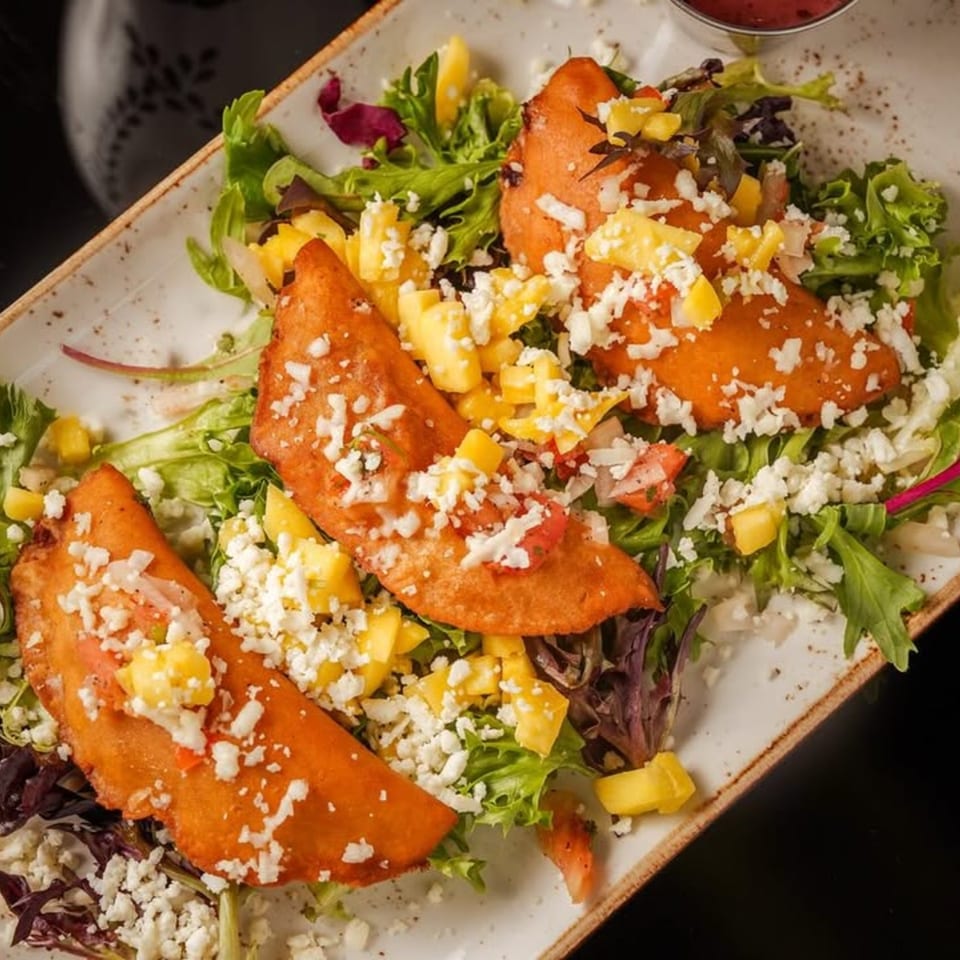 Three Mezcal empanadas on a bed of greens, topped with crumbled cheese and diced mango.